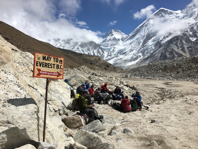 Group of hikers resting with mountains in the background and a sign pointing to Everest Base Camp.