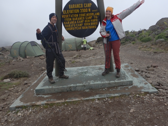       Partial view of two people posing with a camp sign, some tents visible.
  
