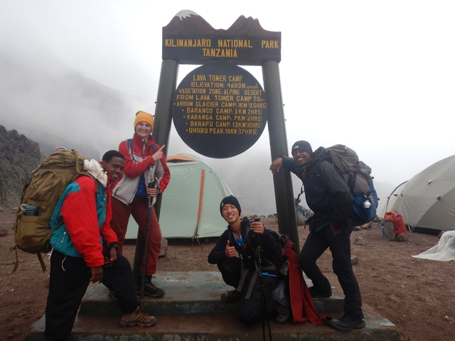       Group of hikers posing with a sign at a camp with tents and cloudy weather.
  