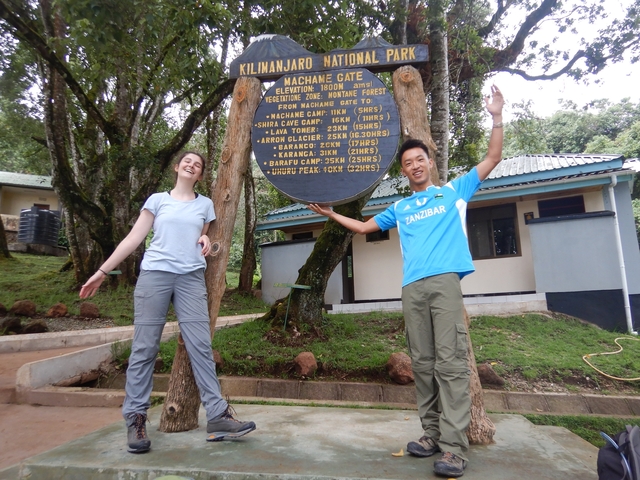      Two people posing at Machame Gate of Kilimanjaro National Park.
  