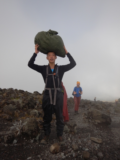       Person carrying a stone on their head during a hike in a foggy, rocky landscape.
  