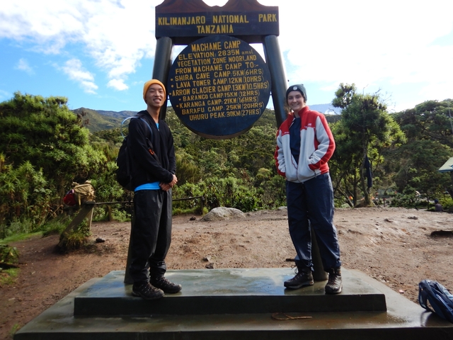       Two people posing at a sign in Kilimanjaro National Park.
  
