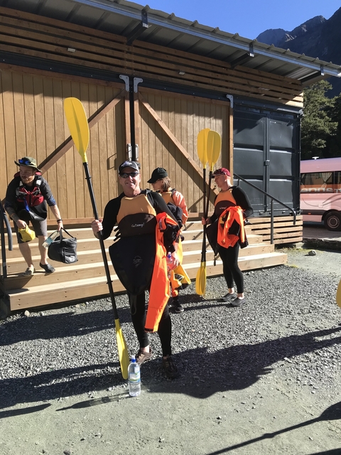       People holding kayaks and paddles outside a wooden building.
  