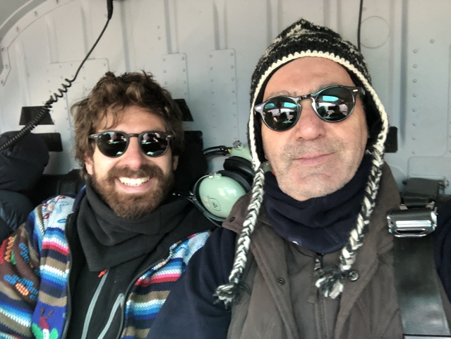       Two men with sunglasses and winter clothing inside a helicopter.
  
