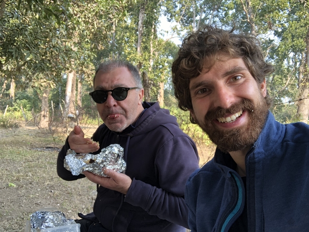       Two men enjoying a meal outdoors in a forest setting.
  