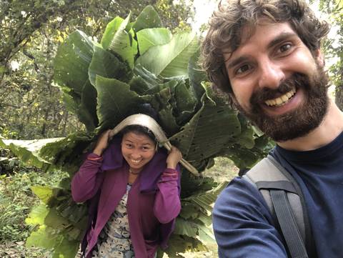       Woman carrying large leaves on her back smiling at the camera.
  