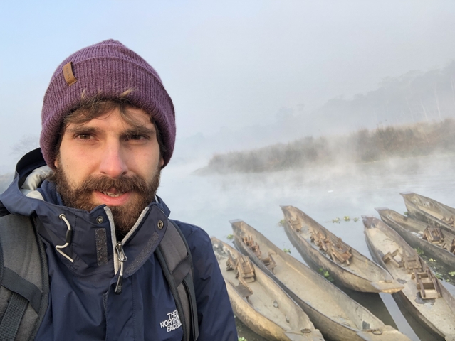      Man taking a selfie near wooden boats on a misty river.
  