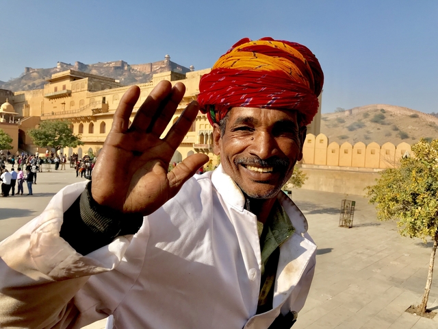       Man in traditional attire smiling and waving with fort in the background.
  