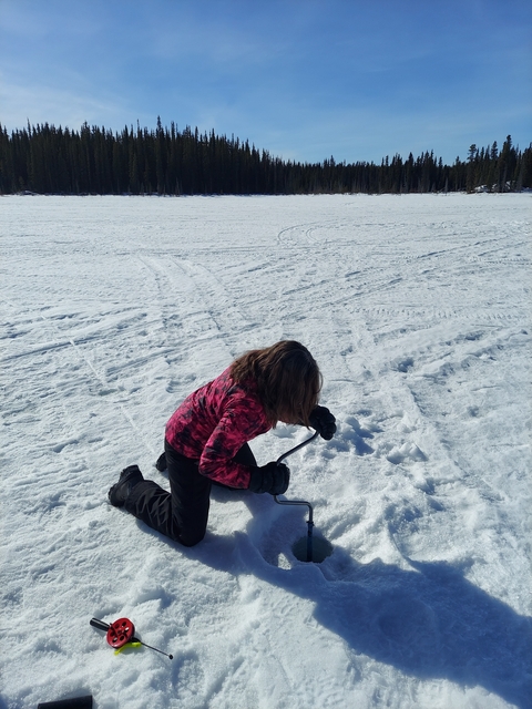 Person in red jacket kneeling on snow with an ice pick.