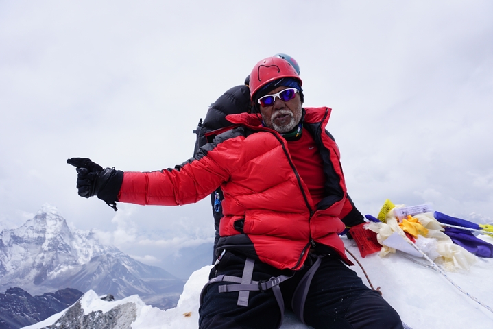 A climber sitting on a snowy peak pointing at the horizon.