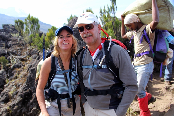 Two hikers smiling on a rocky trail with a porter in the background.