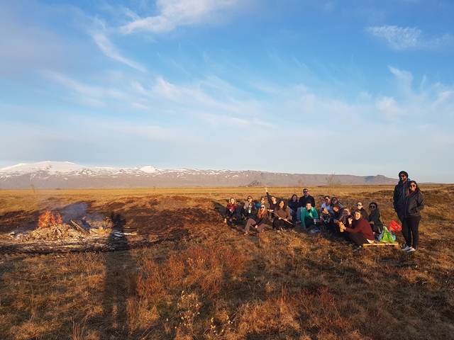       Group of people sitting by a bonfire on an open plain with mountains in the background.
  
