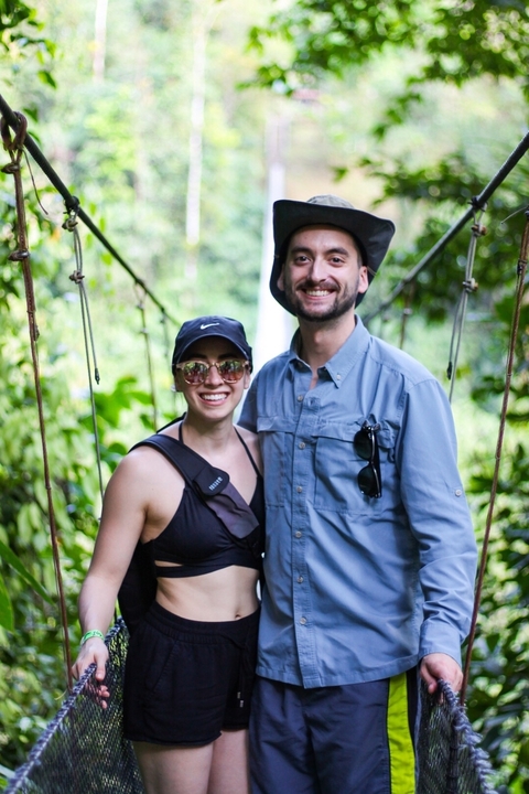 Couple smiling on a suspension bridge in a forest.