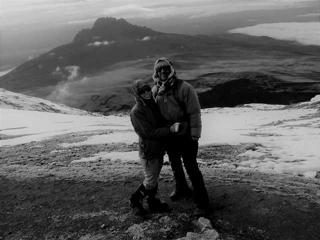       Two people in winter clothing standing on a snow-covered mountain with a scenic view.
  