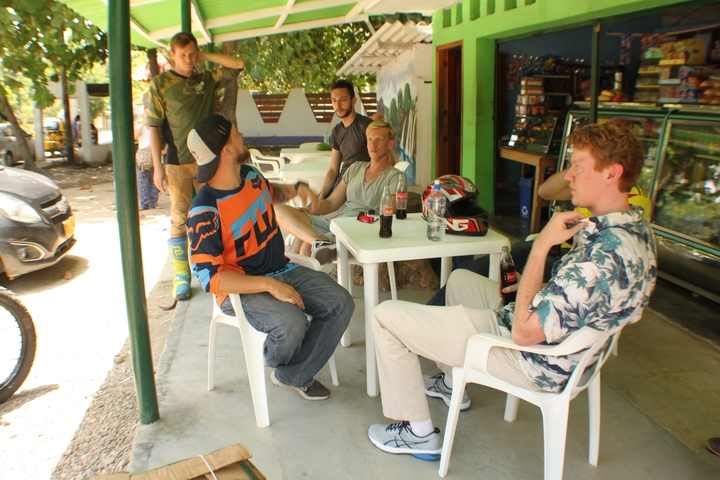       Group of people seated around a table outside a shop, with motorcycling gear visible.
  