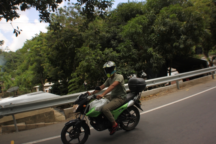       Person on a motorcycle wearing a helmet on a road surrounded by greenery.
  