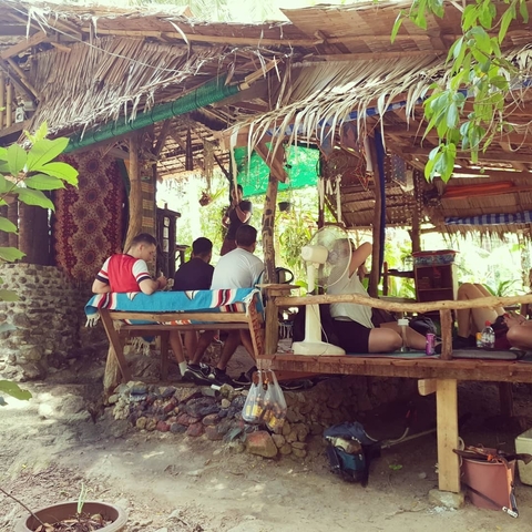 People relaxing in a rustic open-air setting with trees and wooden furniture.