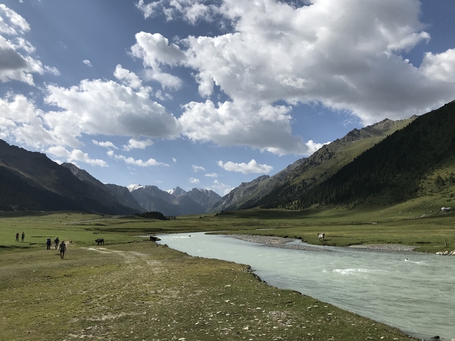 People walking along a river with mountains in the background