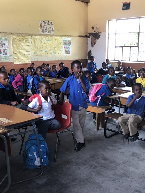       Children in a classroom sitting on chairs and benches.
  