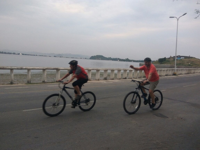       Two people cycling on a road by a lake.
  