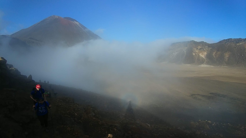 Hikers on a rocky path with a volcanic peak partly shrouded by clouds.