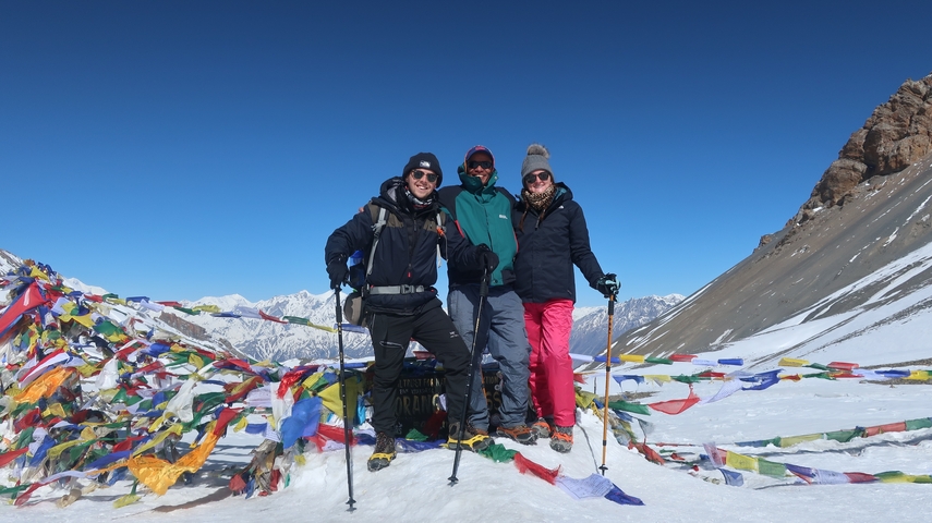       Three people posing at a snowy pass with prayer flags.
  