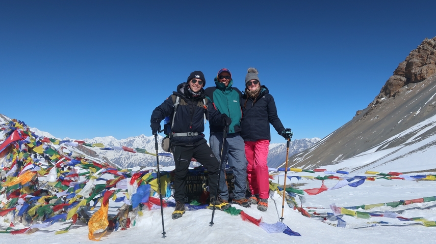       Three people posing at a snowy mountain pass with prayer flags.
  