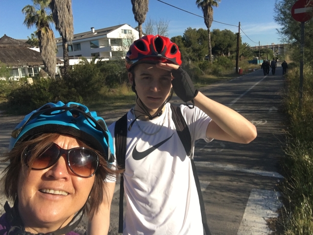       Two people with bicycles on a road near trees and buildings.
  