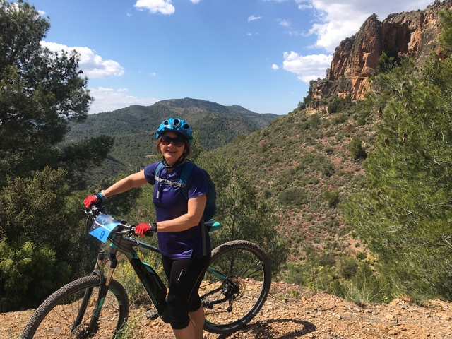       Woman with a bicycle, posing outdoors with mountains in the background.
  