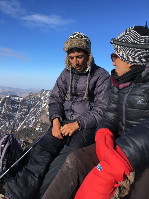 Two people dressed for cold weather sitting outdoors with mountains in the background.