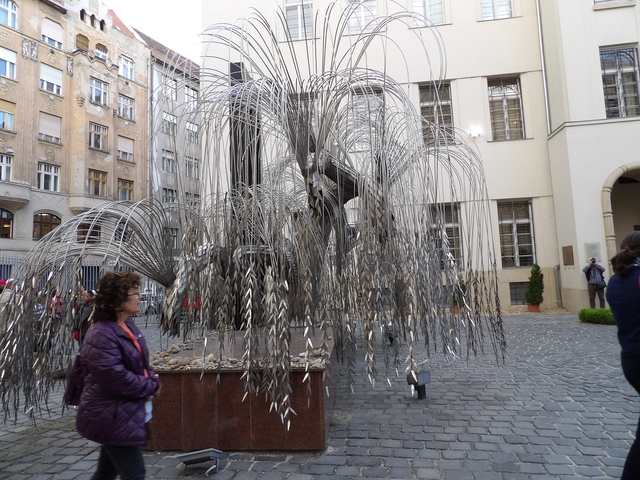       Statue of a weeping willow with metallic leaves and people nearby.
  