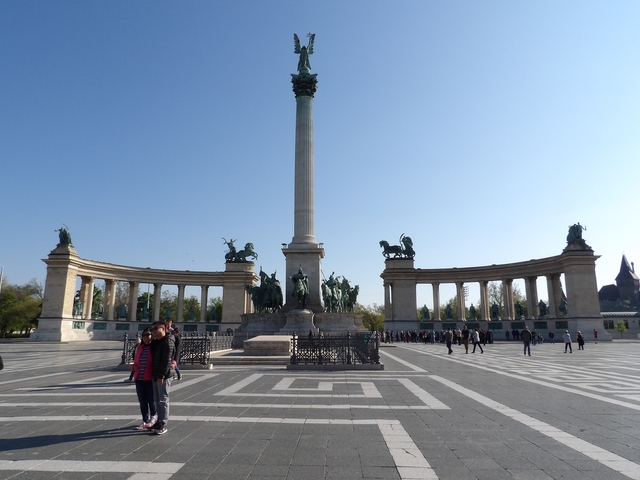 Historic square with statues, trees, and visitors.