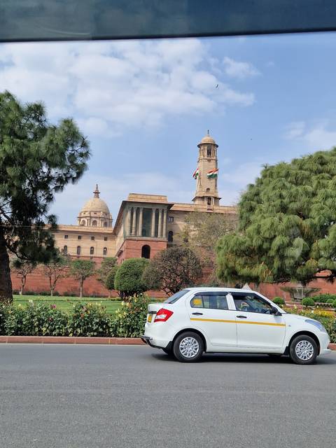       Government building with dome and flying Indian flags viewed from a moving car.
  