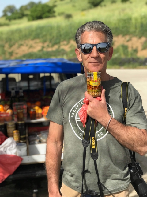       Man holding a jar of preserved fruit near a market stand.
  