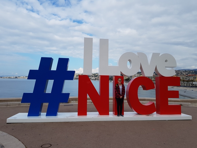 Person standing in front of a large sign that says 'I Love Nice' by the beach.
