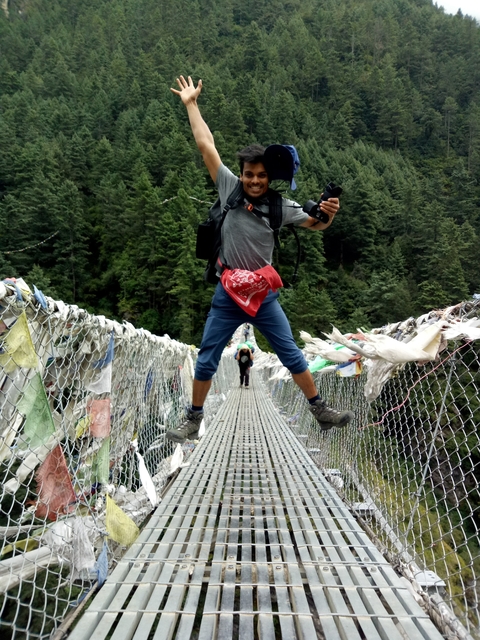 Person jumping on a suspended bridge with prayer flags.