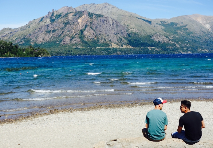      Two people sitting by a lake with mountains in the background.
  