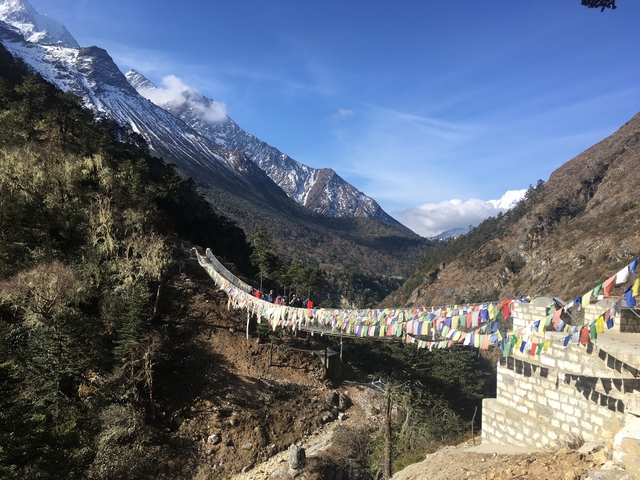      Bridge with colorful prayer flags and hikers in a mountainous landscape.
  
