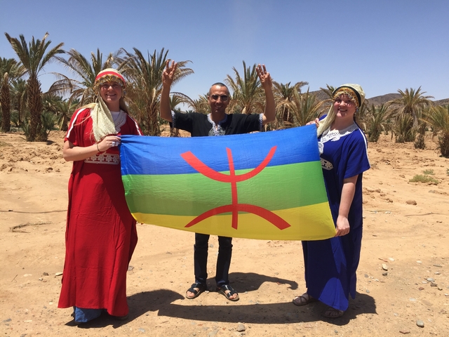Three people holding a colorful flag in a palm grove.