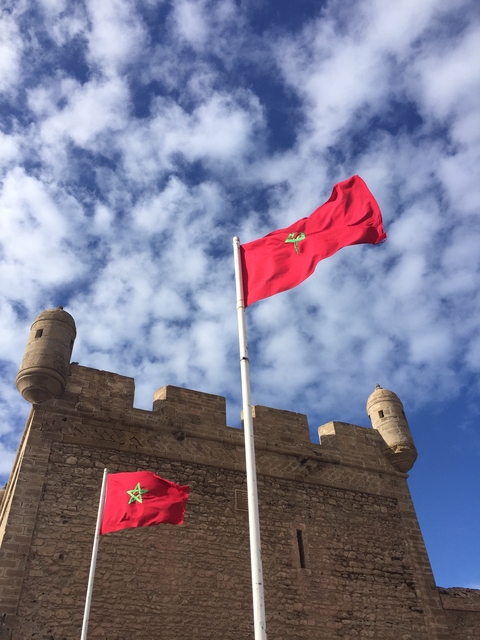       Moroccan flag waving atop a fortified wall under a blue sky.
  