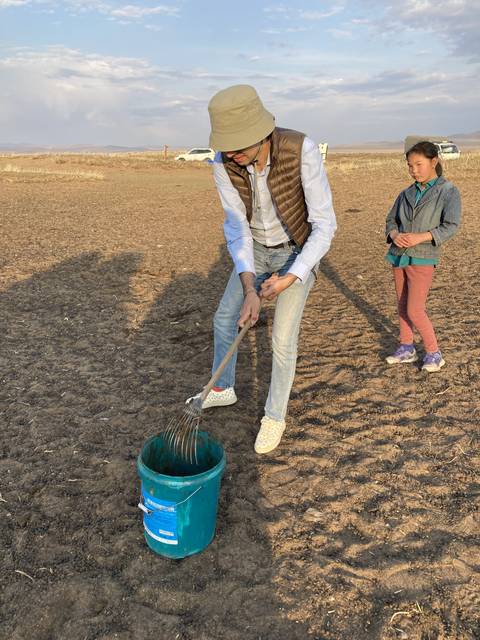 Person scraping dirt into a blue bucket in an open field.