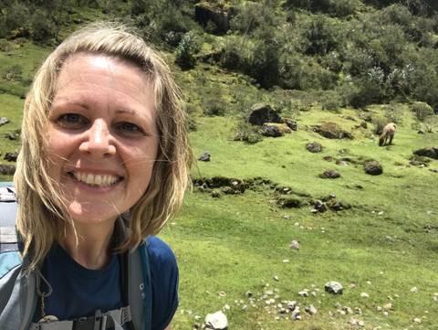 A person smiling with a backpack in a grassy area with a horse in the background.