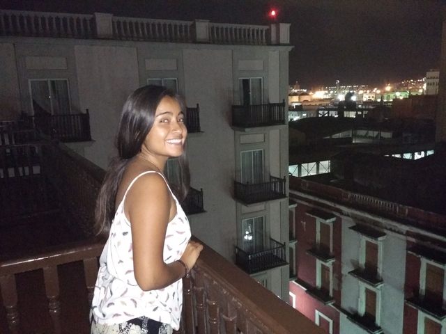       A woman smiling on a balcony against a night city skyline.
  