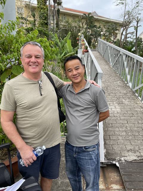       Two people posing on a bridge with greenery.
  