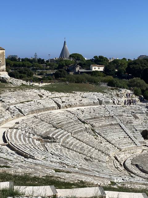       Ancient amphitheater with stone seating and modern buildings in the distance.
  