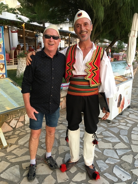 Two men posing together, one in traditional attire, in front of a shop.