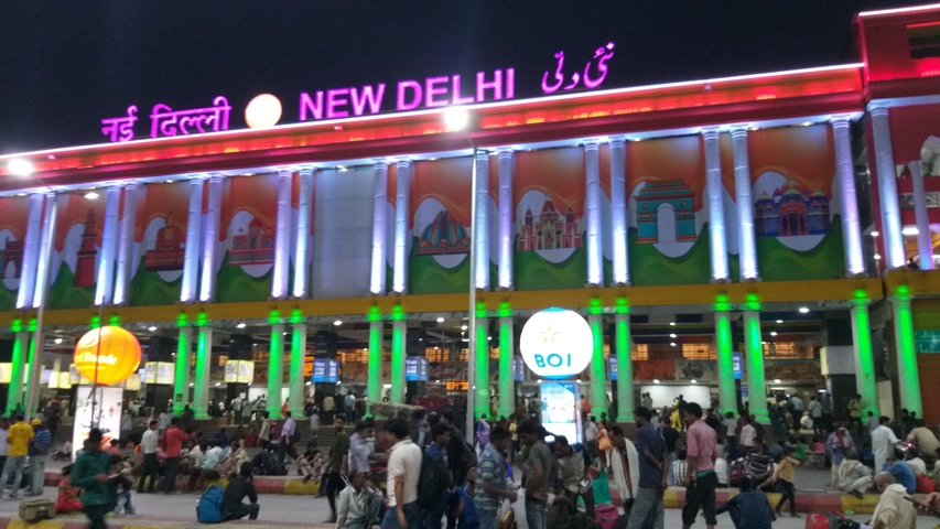       Night view of New Delhi railway station with vibrant lights and people gathering outside.
  