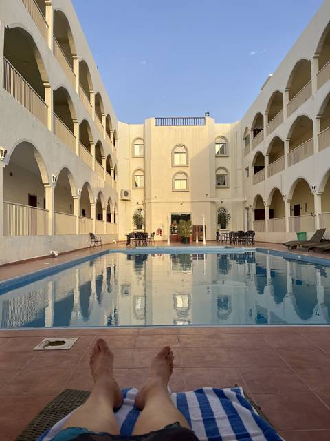       View of a swimming pool with a hotel building and a person relaxing with feet visible.
  