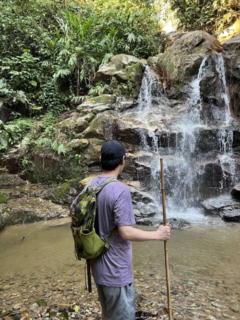 Person standing with a walking stick near a small waterfall.