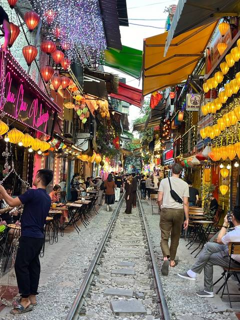       Street with railroad tracks and colorful lanterns, bustling with people.
  